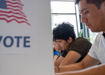 Hispanic young man is seen intently casting his vote in a U.S. election. The voting booth is adorned with an American flag and the word "VOTE” democracy, voting rights, and electoral participation.